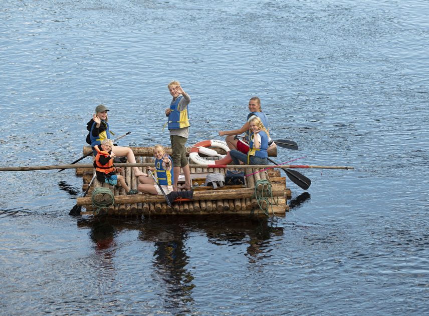 Mitt på älven och omgiven av glittrande vatten flyter en liten timmerflotte utan tältkonstruktion i Klarälven. Ombord på flotten syns många människor som avbryter sin paddling för att vinka till fotografen.