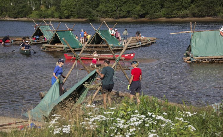 Ett nästan färdigt timmerflottsbygge i Klarälven. De flesta av flottarna har redan sin tältkonstruktion färdig, i förgrund lägger en grupp människor presenningen som ett tak över stockarna.