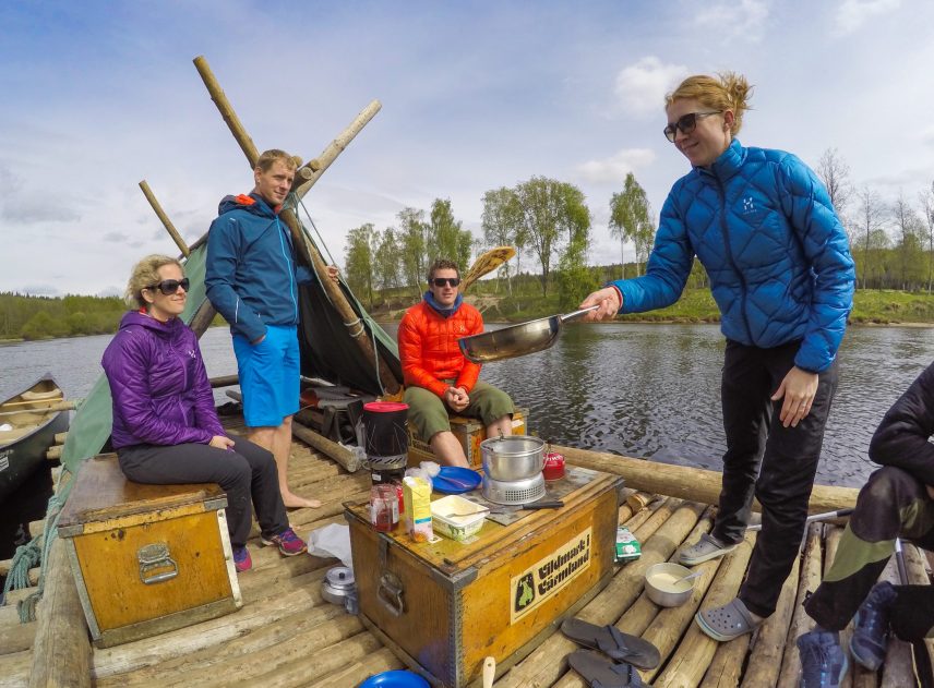 Matlagning ombord på en flotte medan den flyter på Klarälven. Fyra personer syns till, dels stående, dels sittande på trälådor. En låda är dukat som bord. En kvinna vänder en pannkaka i luften och fångar den med en stekpanna. Soligt väder.