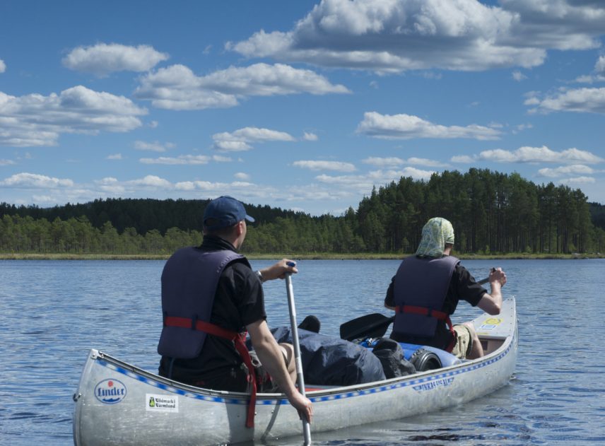 Två personer sitter i en kanot som även är lastad med mycket packning. De är på väg till motsatt sida sjön där en kulle täckt av skog tornar upp sig. Människorna skyddar huvuden mot solen men himlen börjar bli molnig.