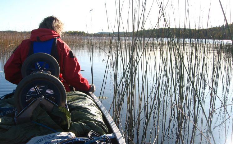 I en fullastad kanot sitter en kvinna i röd jacka. Högst upp på packningen ligger en uppfälld men upp och ner vänd kanotvagn. Kanoten ligger i vattnet i en spegelblank sjö, direkt vid kanotens sida sticker stort sjögräs upp ur vattnet.