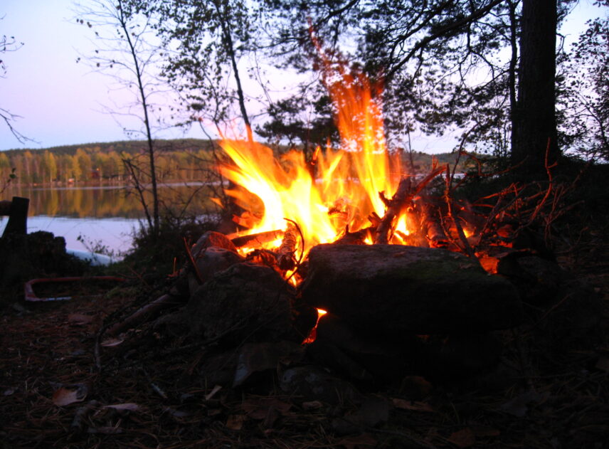 En kraftigt brinnande brasa dominerar hela bilden. Eldstaden bildas av stora svarta stenar. Bakom lågorna skyms en sjö som ligger ljust i kvällssolen.