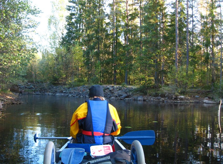 På framsätet i en kanot sitter en person med gul jacka och blå flytväst. Hon syns bakifrån. Hon har en paddel i knän men paddlar inte. Det är ett mindre vattendrag. Granar och björkar rader upp sig längs stranden.