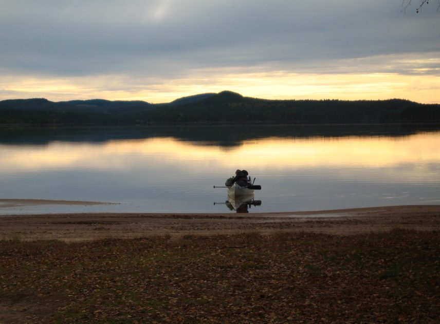 En fullpackad kanot ligger redo för avfärd vid stranden. Det är tidig morgon, en gul remsa ljus syns bakom den svarta skogen på andra sidan sjön och speglas i det stilla vattnet. Himlen har inte hunnit bli blå än, nattens grå moln är kvar.