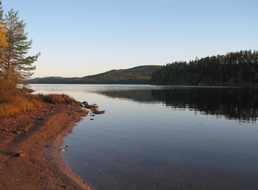 En stenig strandlinje begränsar en spegelblank sjö. På andra sidan vattet skär en skogsklädd bergskedjan genom bilden. På en ljusblå himmel syns inte ett enda moln.