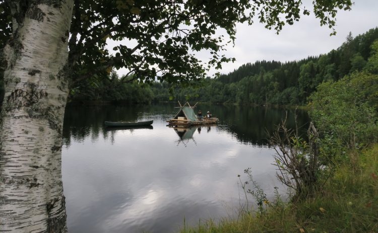 Bilden är tagen från land och ramas in av Klarälvens strandvegetation. Till vänster begränsas fotot av en stor björk. På älven i mitten syns en timmerflotte med tältkonstruktion som släpper en kanot efter sig. Ombord på flotten anas flera personer samt trälådor och annan packning.