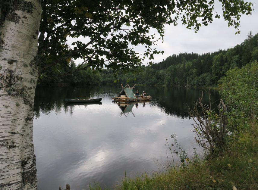 Bilden är tagen från land och ramas in av Klarälvens strandvegetation. Till vänster begränsas fotot av en stor björk. På älven i mitten syns en timmerflotte med tältkonstruktion som släpper en kanot efter sig. Ombord på flotten anas flera personer samt trälådor och annan packning.