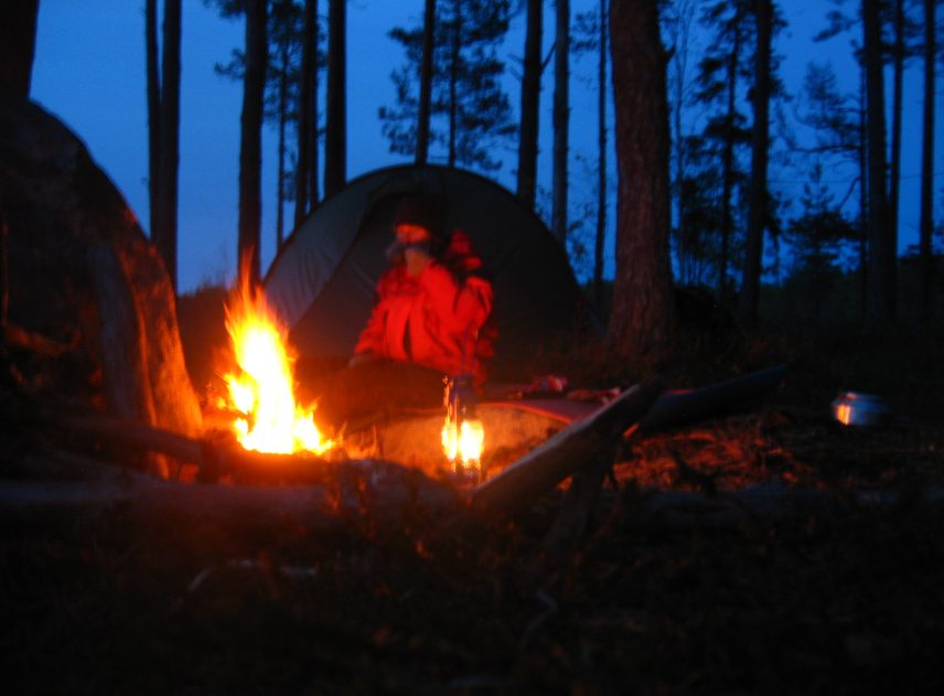 En brasa i förgrunden är bildens enda ljuskälla. Bakom den syns ett tält och en person i röd jacka som värmer sig vid elden. Den glesa skogen ger blicken fri på en mörkblå himmel.