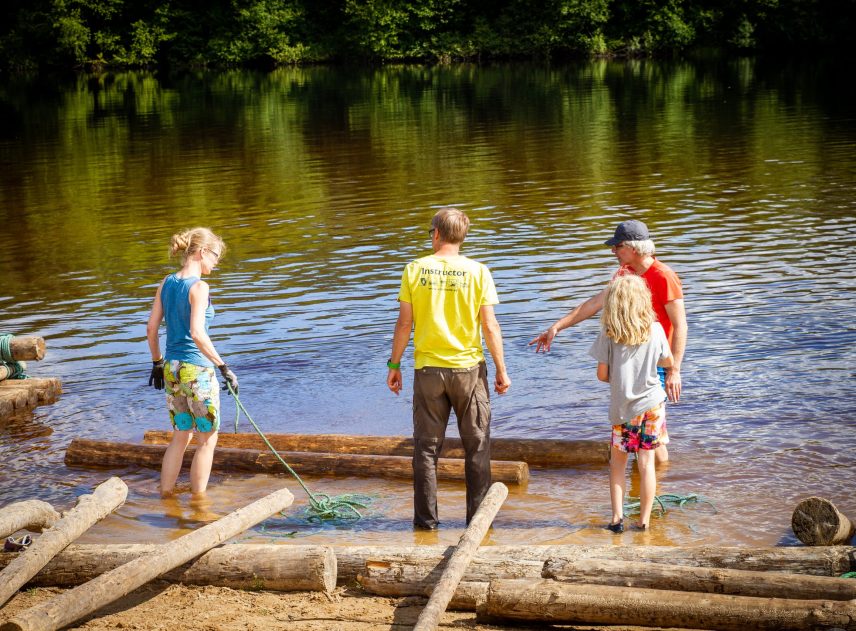 Ett bygge av en timmerflotte har precis börjat. Fyra personer står i vattenbrynet och ska binda ihop stockarna med ett rep. Klarälvens vatten speglar grönskan från växtligheten på andra strandkanten.