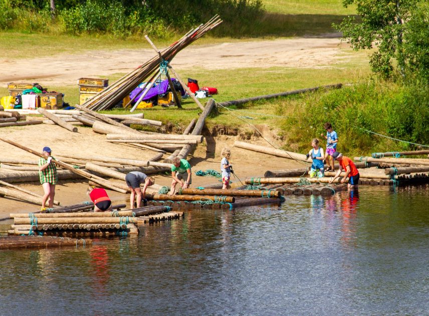 Byge av timmerflottar vid Klarälvens strand i Branäs. Personer i vatten och på land, halvfärdiga flottar ligger i vattnet. Det verkar vara en solig dag.