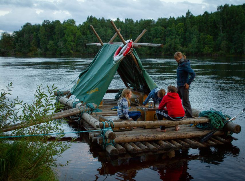 Fyra personer i olika åldrar ombord på en timmerflotte. De står och sitter samlade runt en trälåda som fungerar som bord. De kanske spelar ett spel eller förbereder mat. Flotten är förtöjd på Klarälvens ena strandkant. I bakgrunden syns den motsatta älvsidan med mycket växtlighet.