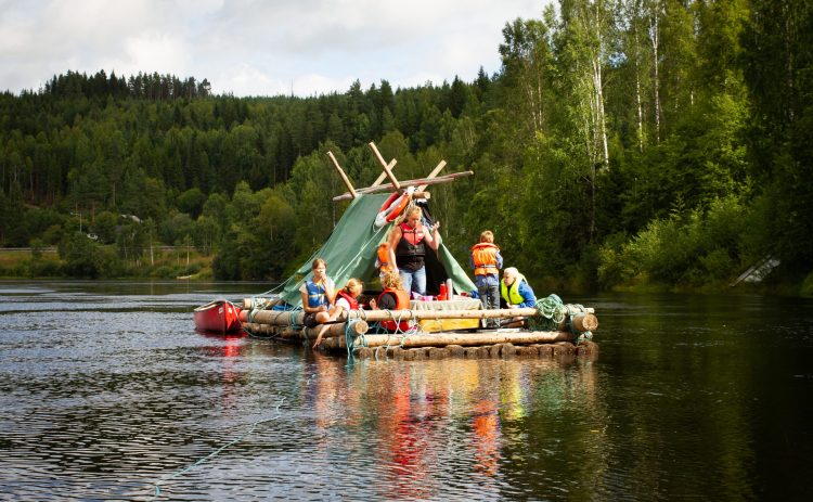 En timmerflotte med tältkonstruktion flyter på Klarälven. Ombord på flotten finns flera personer, de flesta barn. En trälåda i gruppens mitt är dukad som bord, kanske till lunch. I bakgrunden mycket växtlighet.