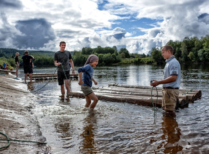 Ett timmerflottsbygge i Klarälven pågår vid Klarälvens Camping. Flera personer står i vattnet, stockarna för flottens första lager ligger redan i älven. En man försöker binda ett rep, en pojke springer genom vattnet för att hjälpa honom.