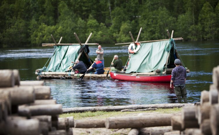 Två stora timmerflottar med tältkonstruktion lämnar byggplatsen. Personerna ombord försöker paddla flottarna ut i Klarälvens huvudströmmen. En pojke står vid stranden och ser efter de försvinnande flottarna. I förgrund syns travar av timmerstockar.