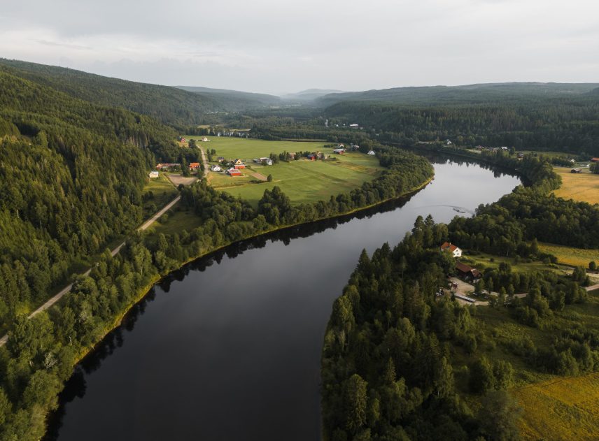 En luftbild visar en av Klarälvens kraftiga meanderbågar. Vid horisonten tornar sig skogsklädda berg upp. Längs älven syns både gårdar med öppna fält och en remsa med mycket träd närmast vattnet.