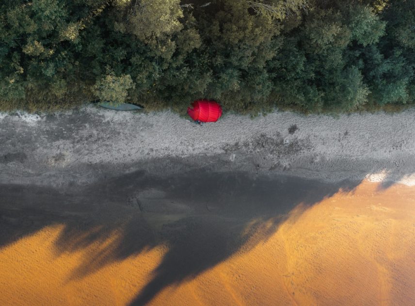 Högt upp ifrån luften syns Klarälvens gula, sandiga botten rakt genom vattnet i bildens nedre del. Den övre delen är täkt med skogens grönska. Däremellan går en smal remsa av ljus sandstrand. Mitt på bilden lyser en röd prick som är ett tält från Hilleberg.
