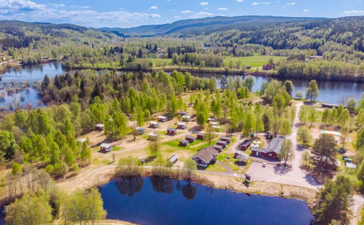 En luftbild visar en campingplats med många stugor. Platsen är på tre sidor omgärdat av vatten, i bakgrunden är det Klarälvens mäktiga meanderbågar, i förgrund syns Korvsjöns djupblå vatten. Ljusgröna blad på träden visar att det fortfarande är tidigt på sommaren.