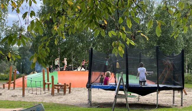 En lekplats med många barn på en studsmatta, en hoppkudde och en hinderbana. Ljusgrön gräsmatta i förgrund, ett träd sträcker sina lövklädda grenar in i bilden. Lekplatsen ligger på Klarälvens Camping.