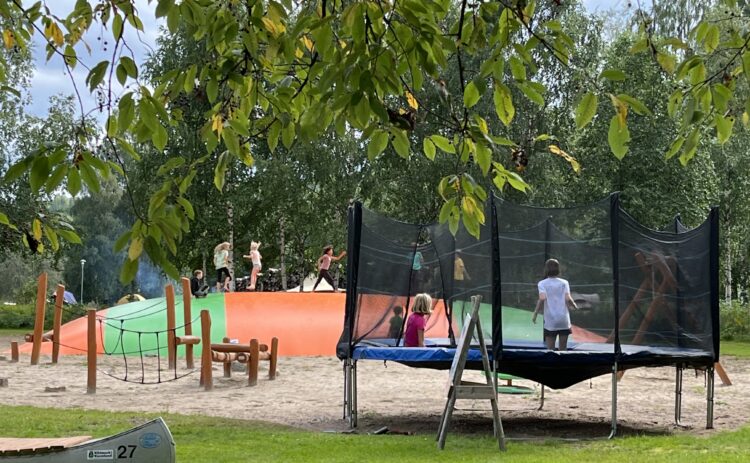 En lekplats med många barn på en studsmatta, en hoppkudde och en hinderbana. Ljusgrön gräsmatta i förgrund, ett träd sträcker sina lövklädda grenar in i bilden. Lekplatsen ligger på Klarälvens Camping.