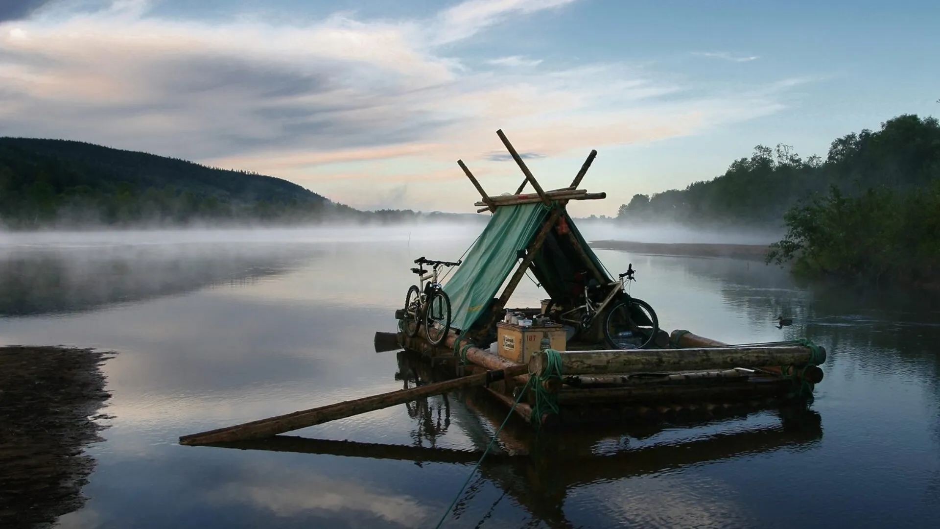 Ett intryck från en flottfärd på Klarälven. En timmerflotte täcker hälften av bilden. Klarälvens vatten ligger i dimma vid horisonten, kanske tidigt på morgonen. På flotten två cyklar, inga människor syns till.